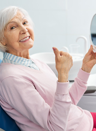 Senior female dental patient giving thumbs up after checking smile in mirror